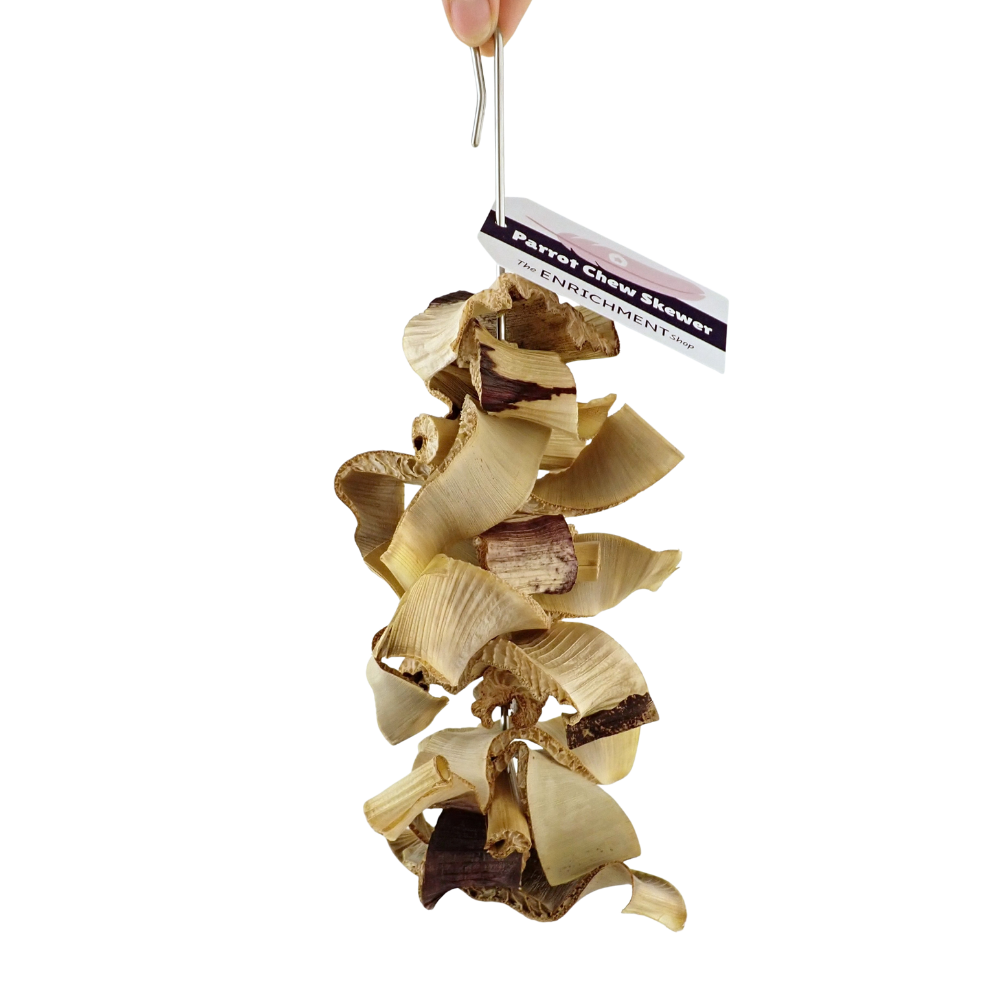 A hanging dog chew toy made of curled wood shavings and bark pieces, suspended from a metal hook with a product tag attached. A human hand is visible holding the hook at the top edge against a plain white background.