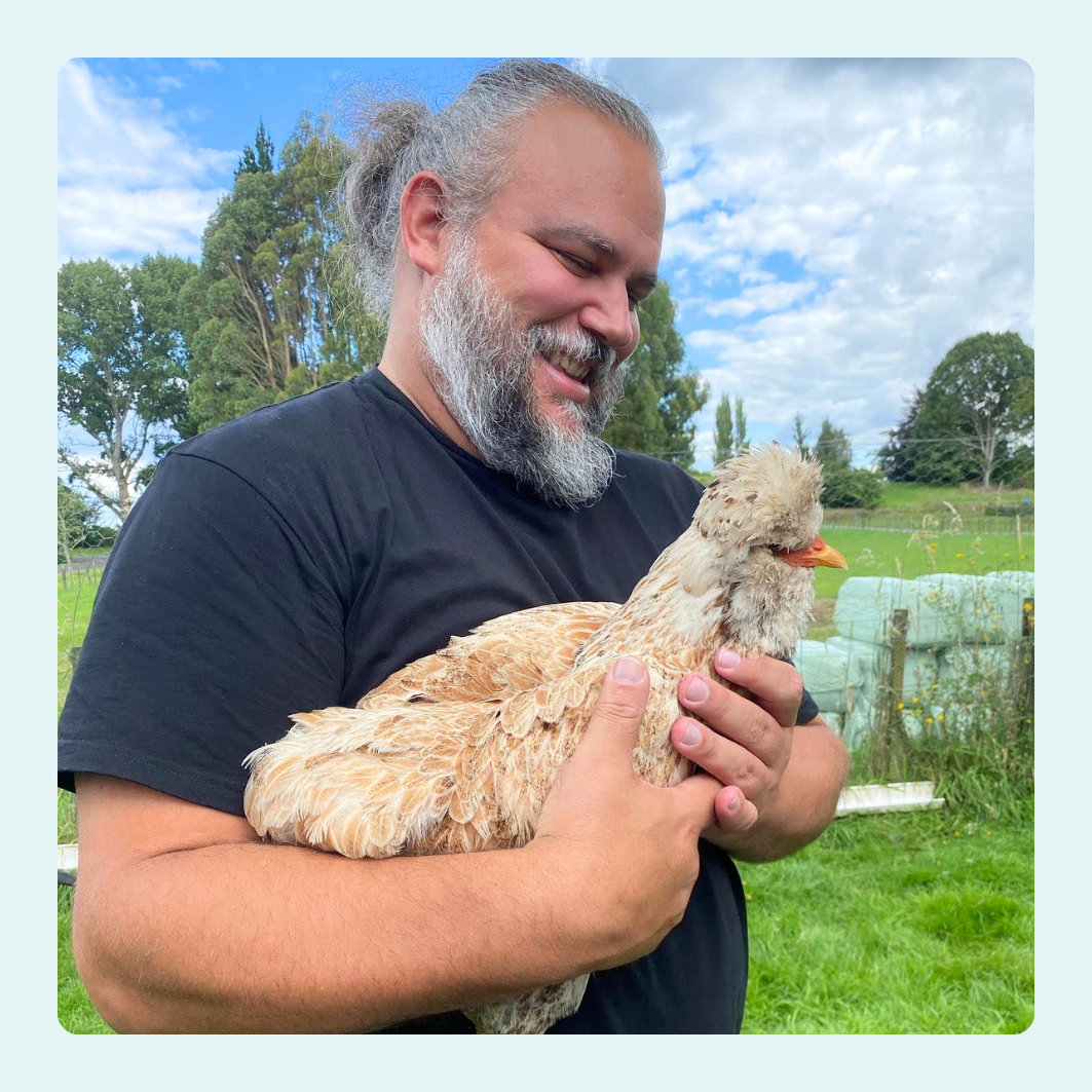 Man holding a chicken outdoors with a grassy field and trees in the background