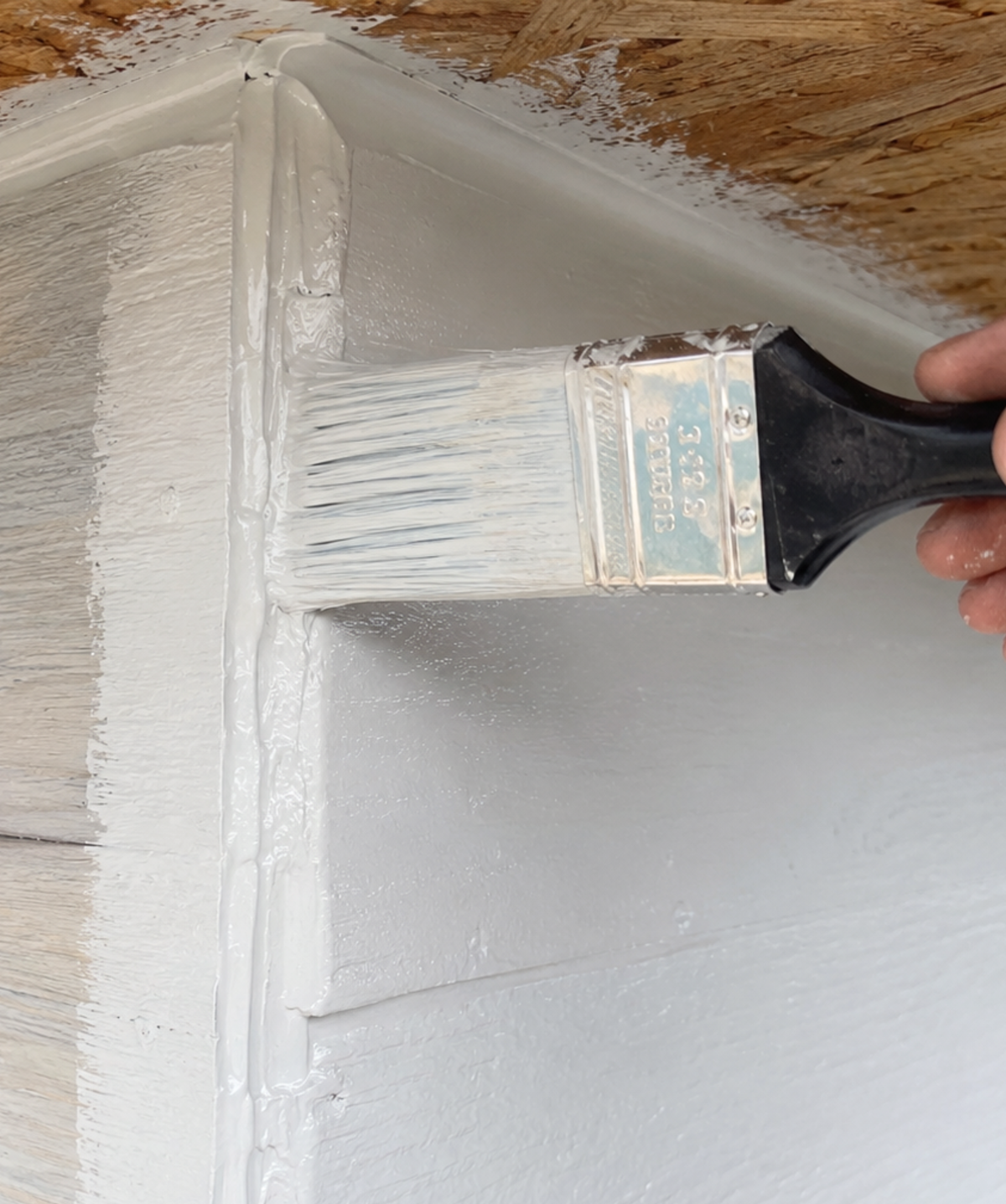 A close-up of a hand holding a paint brush and applying white paint to a wooden surface, with a partially painted corner visible.